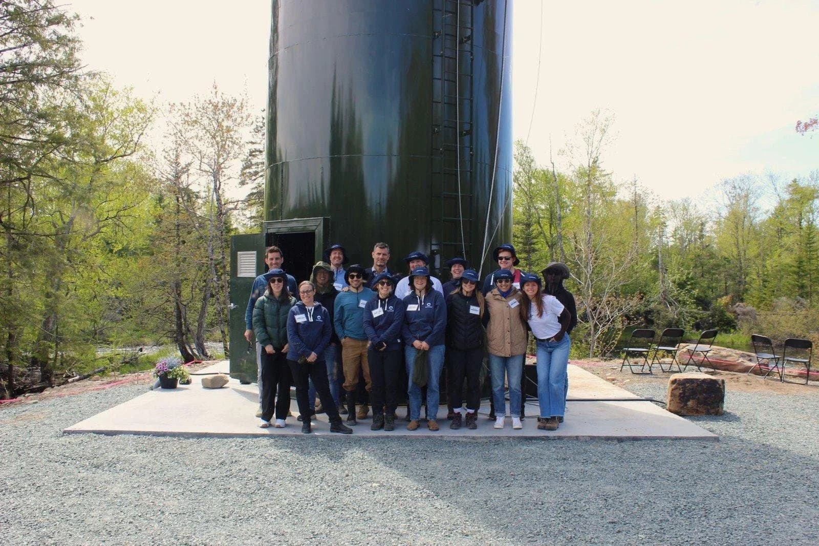 Team members gathered at the base of a tall green dosing silo beside a forested river