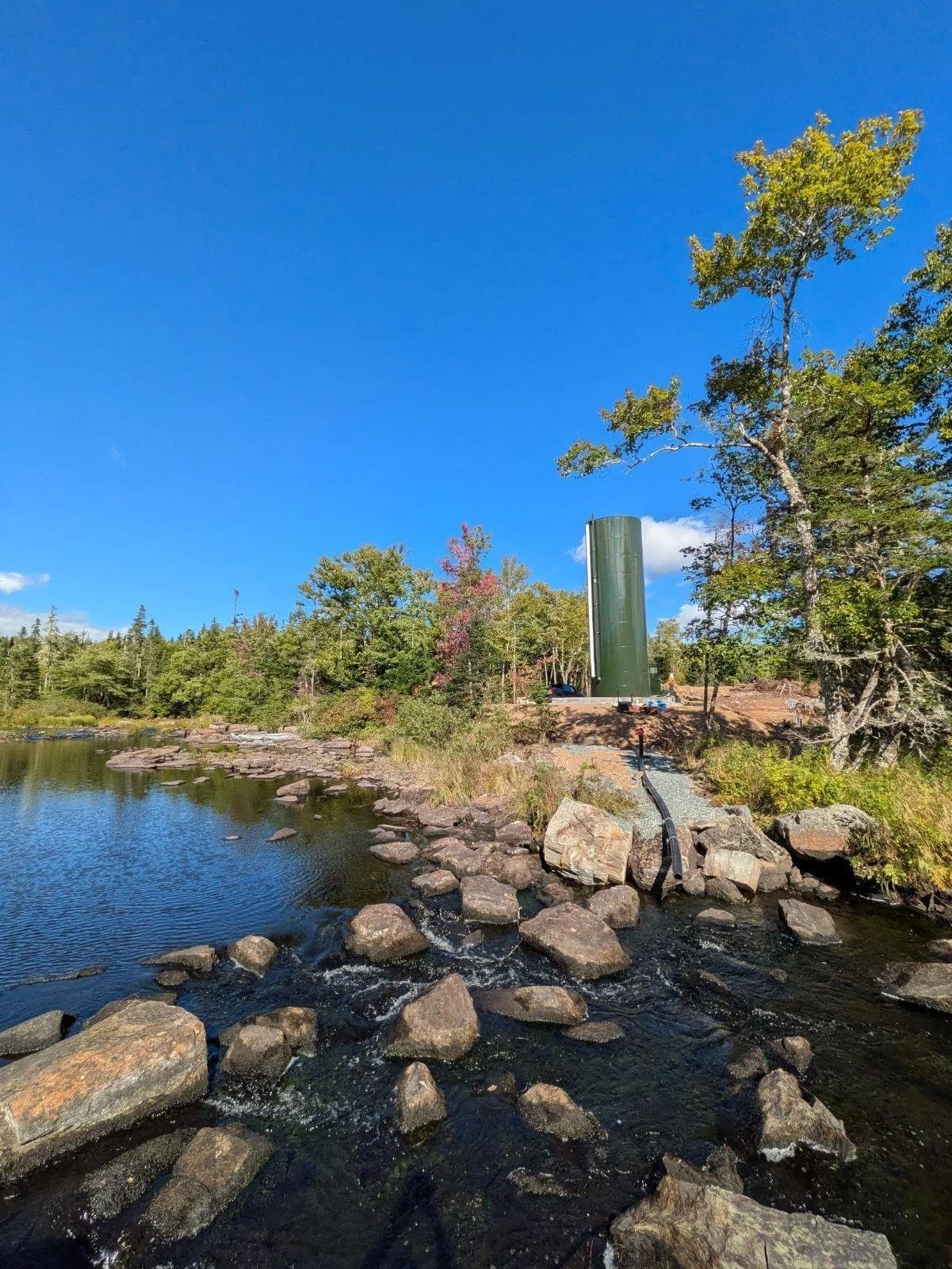 Green dosing silo standing among trees beside a rocky river under a blue sky